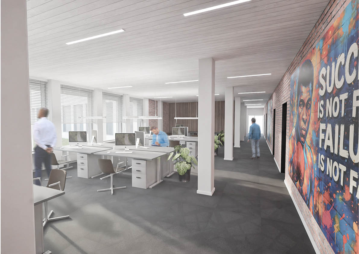 Åbent kontorlokale med flere skriveborde og computere, et par mennesker, der arbejder, store vinduer og et farverigt vægmaleri med motiverende tekst på en murstensvæg.