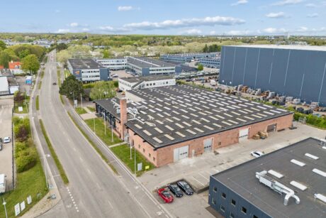 Luftfoto af et industriområde med et stort lagerhus i mursten, en tilstødende parkeringsplads fyldt med biler samt flere omkringliggende kontor- og lagerbygninger.