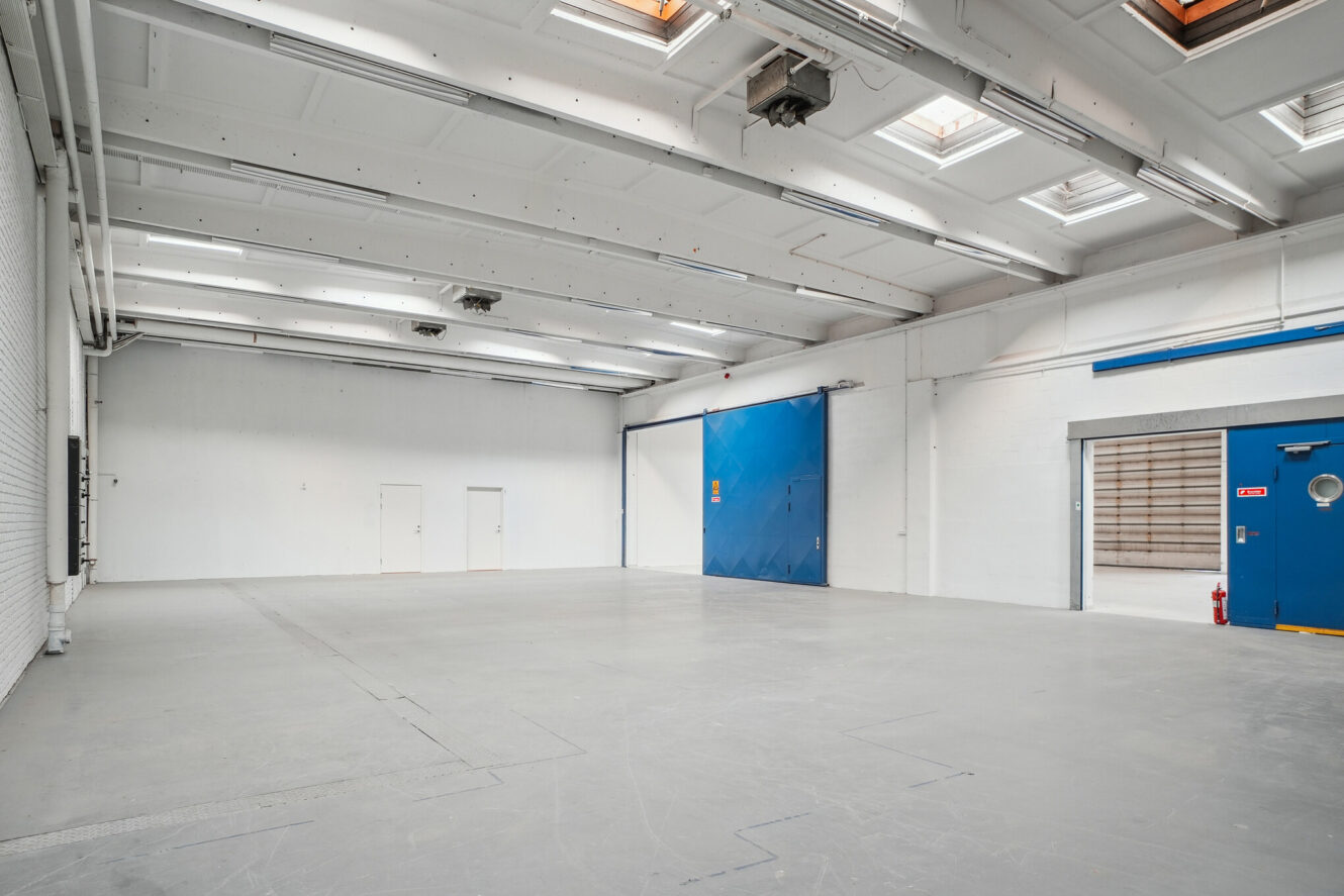 Stort, tomt industrilager med hvide vægge, højt til loftet, ovenlys og blå døre. Poleret gråt betongulv og synlig ovenlys.