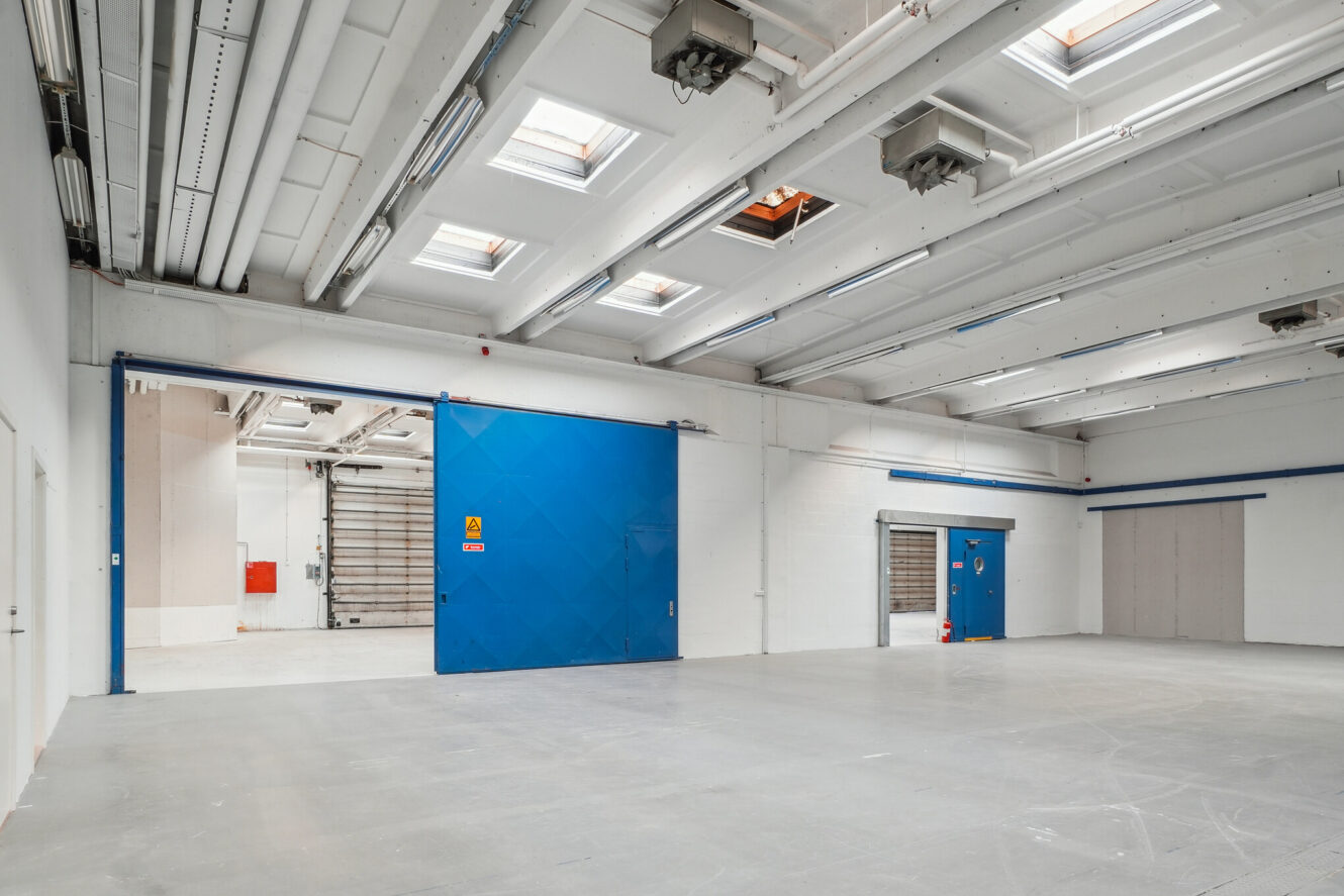 Stort, tomt industrilager med hvide vægge, gråt gulv, ovenlys og store blå skydedøre.