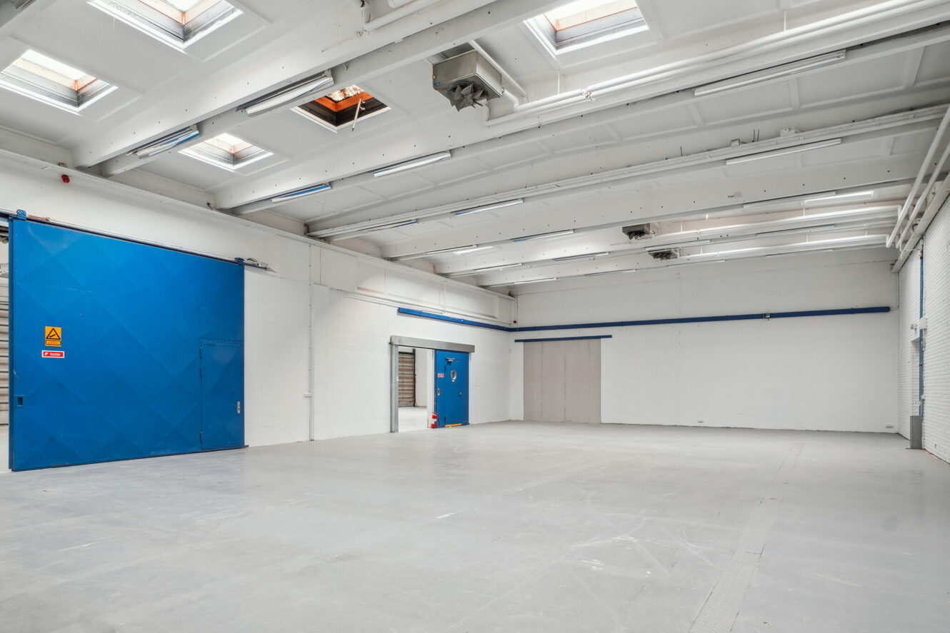 Stort tomt industrilager med hvide vægge, højt til loftet, ovenlys og store blå skydedøre.