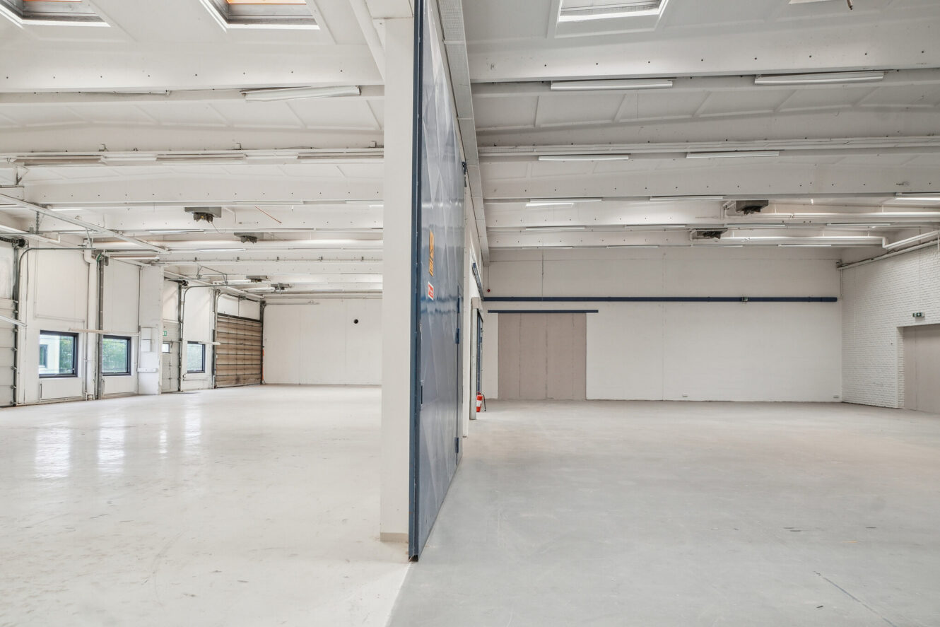 Bredt udsyn over et tomt industrilager, opdelt af en delvis væg, med højt til loftet, ovenlys og hvide gulve og vægge.