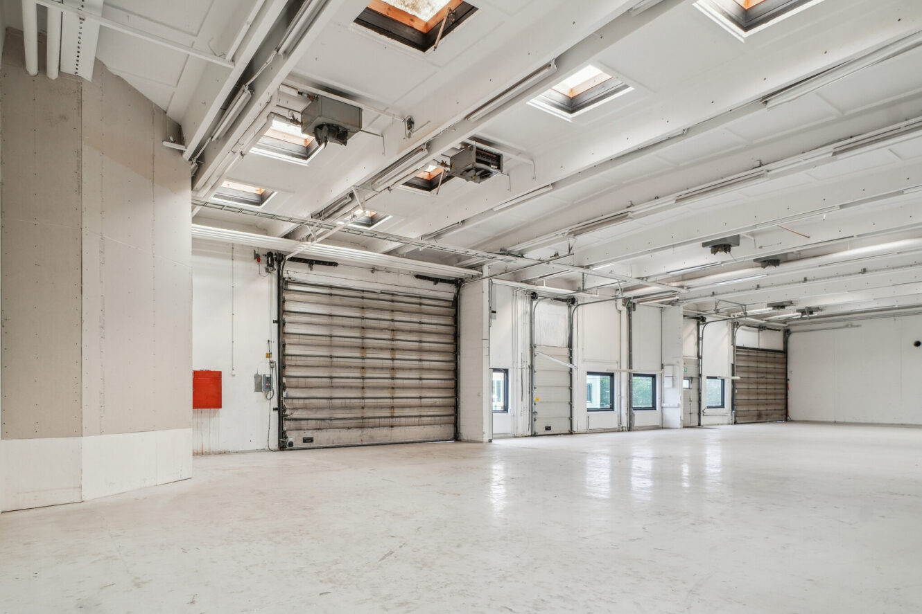 Stort, tomt industrilager med hvide vægge, store overliggende døre, ovenlysvinduer i loftet og synlige rør.