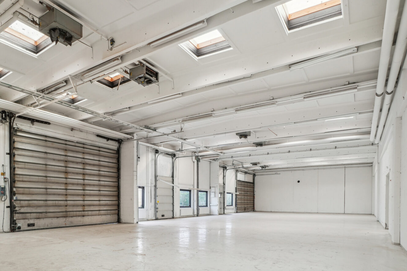 Rummeligt, tomt industrilager med hvide vægge, højt til loftet, ovenlysvinduer, store garageporte og små vinduer, der lukker naturligt lys ind.
