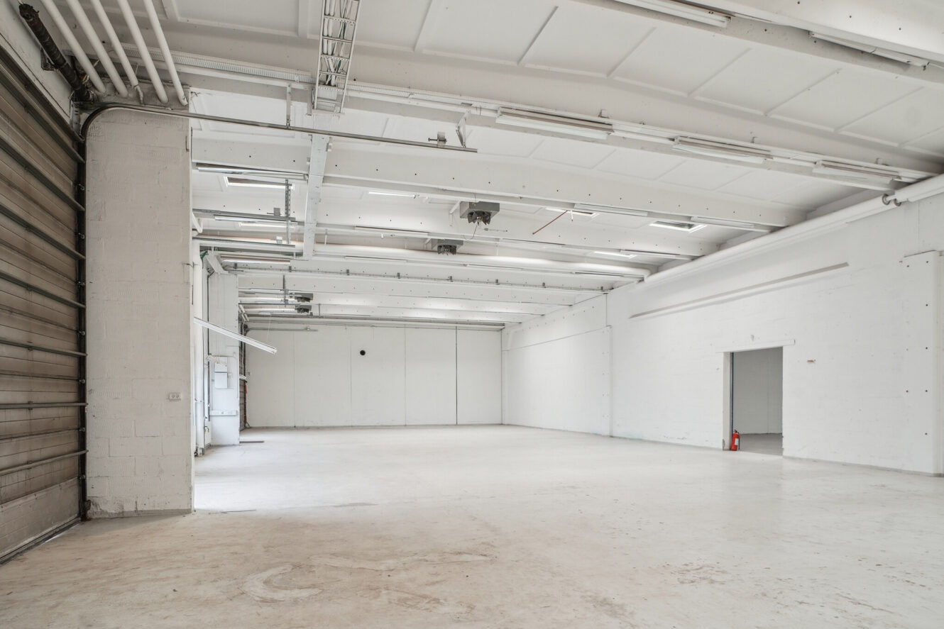 Stort tomt industrilager med hvide vægge, synlige rør i loftet, en sammenrullet metaldør og en enkelt brandslukker i nærheden af en åben døråbning.