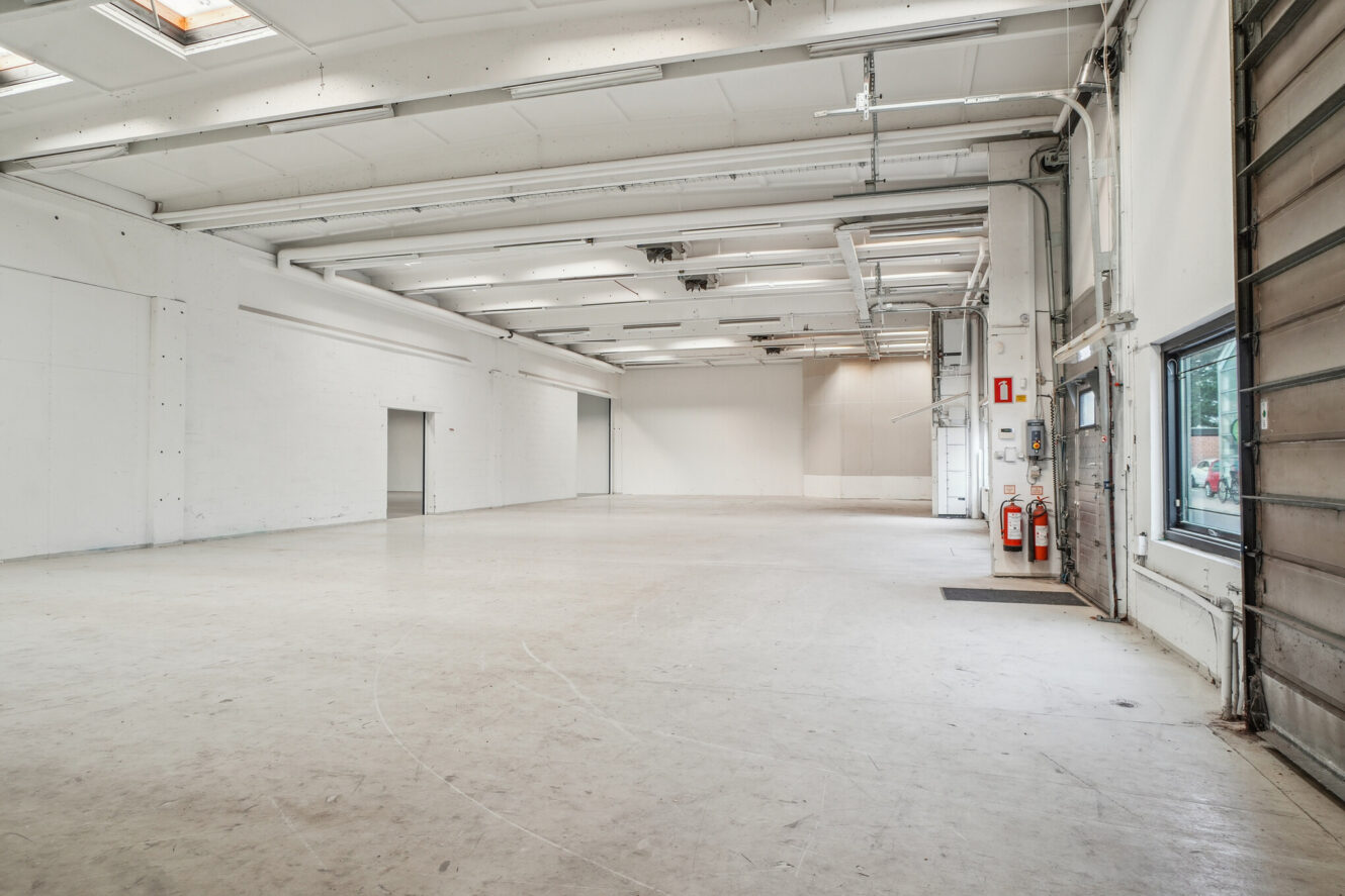 Stort, tomt industrilager med hvide vægge, højt til loftet, synlige rør, brandslukkere og en stor garageport til højre.