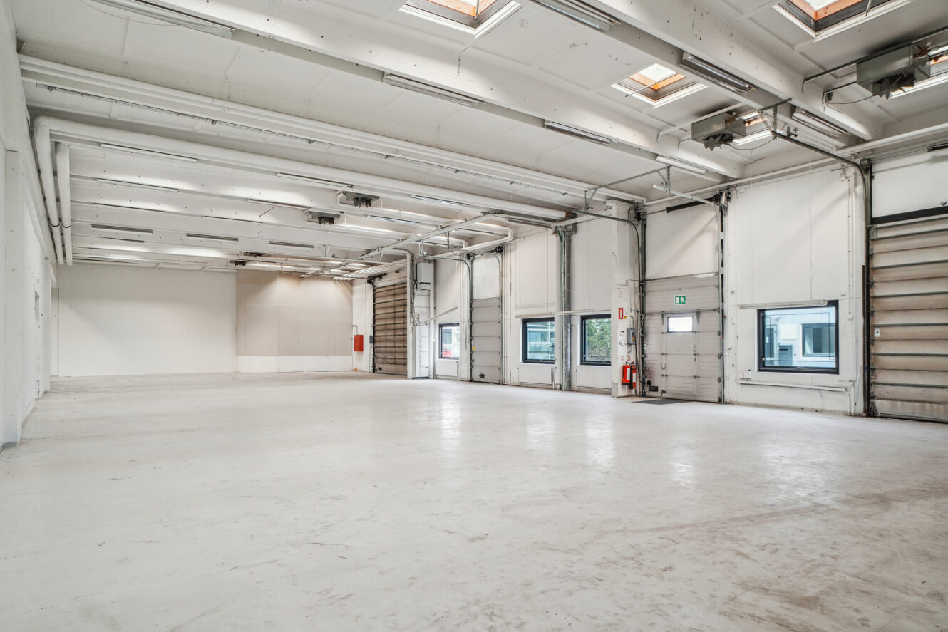 Tomt industrilager med hvide vægge, højt til loftet, ovenlys, flere garageporte og flere vinduer langs den ene væg.