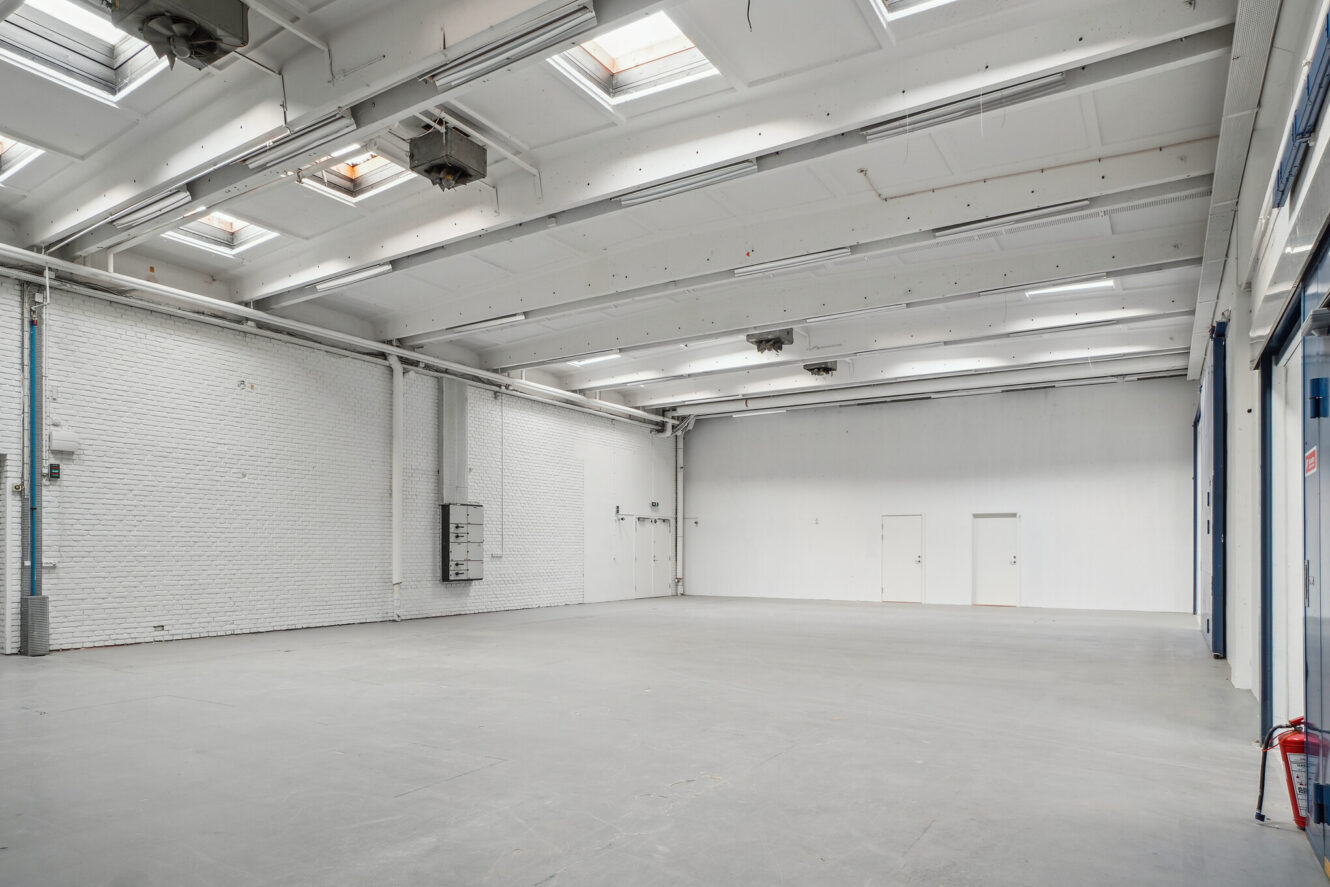 Stort, tomt industrilager med hvide vægge, højt til loftet, ovenlys og betongulv; brandslukker ved glasdør til højre.
