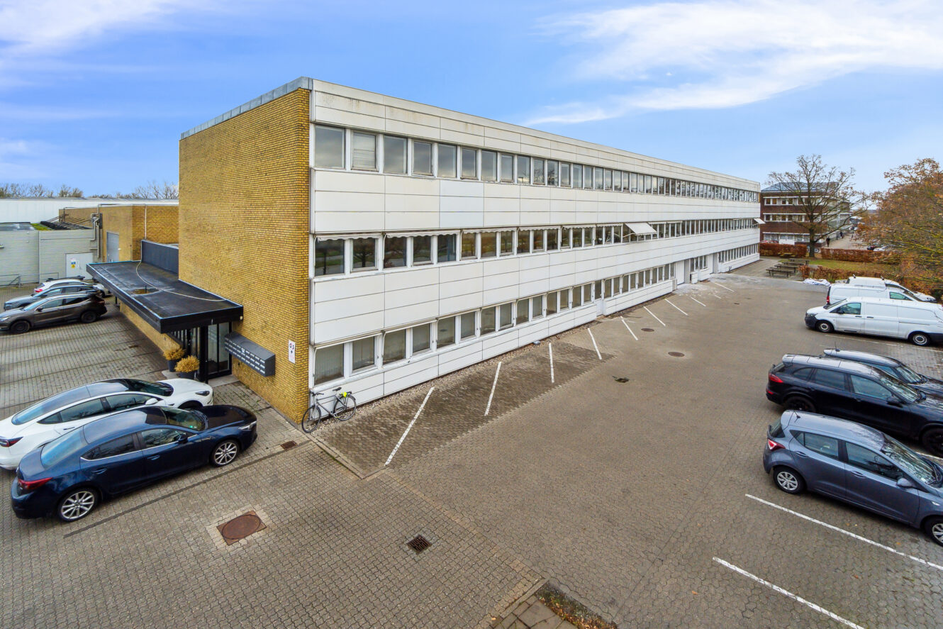 Treetagers kontorbygning med hvid og gul mursten udvendigt, tilstødende parkeringsplads med flere parkerede biler og klar himmel i baggrunden.