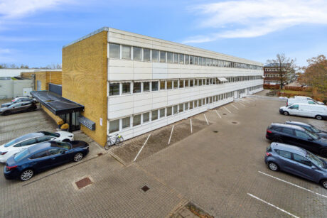 Treetagers kontorbygning med hvid og gul mursten udvendigt, tilstødende parkeringsplads med flere parkerede biler og klar himmel i baggrunden.