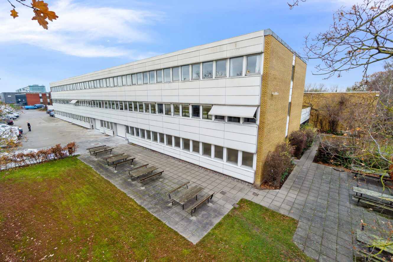 Rektangulær treetagers bygning med mange vinduer, der støder op til et udendørs område med picnicborde og omgivende grønt under en delvist overskyet himmel.