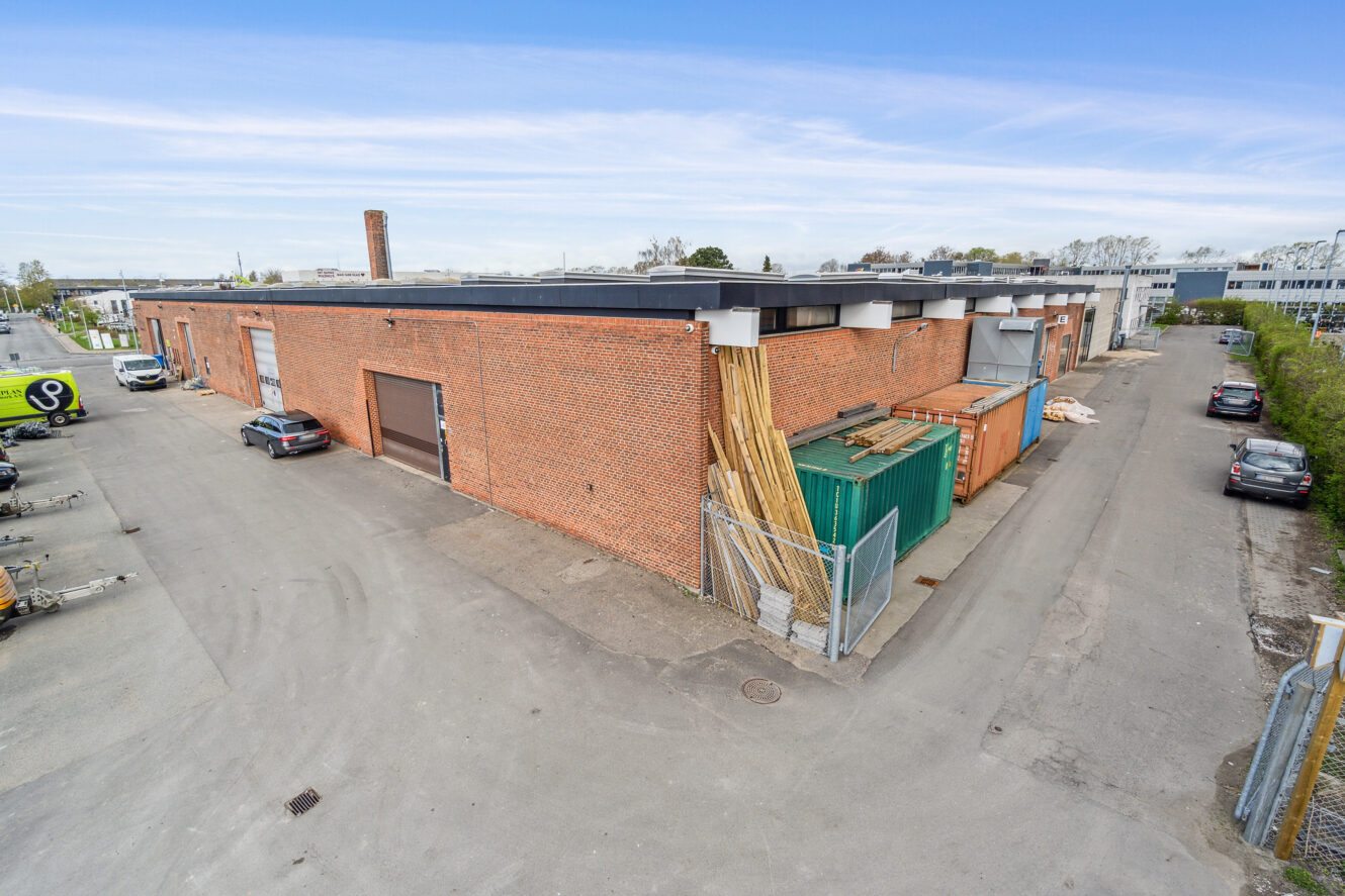 Stort muret industrilager med flere garageporte, indhegnet område med stablet træ og parkerede biler langs en tilstødende vej under en klar himmel.