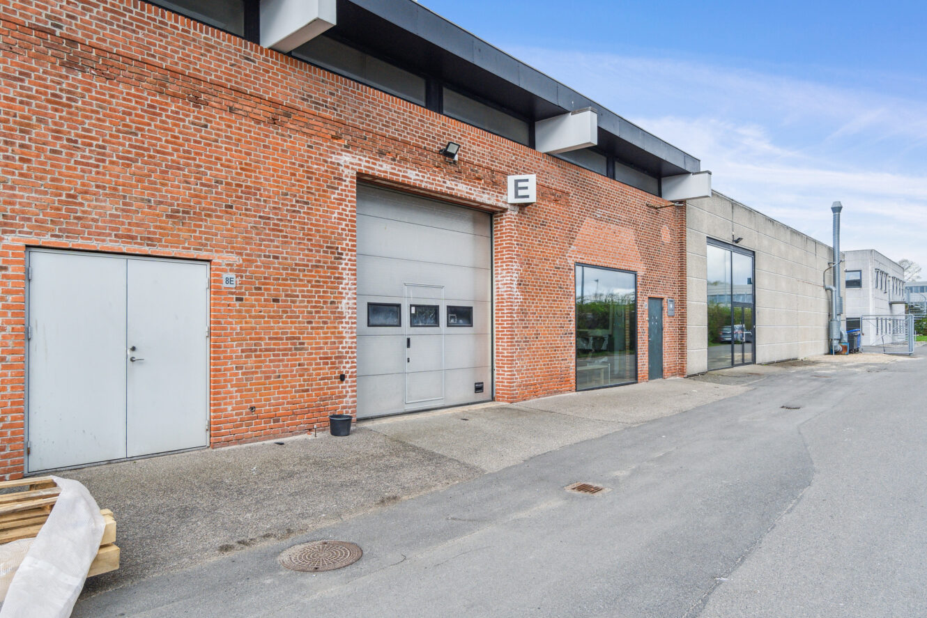 Udvendigt billede af en industribygning med rød murstensfacade, stor garageport mærket E, metaldør, glasvinduer og betonfortov under en blå himmel.