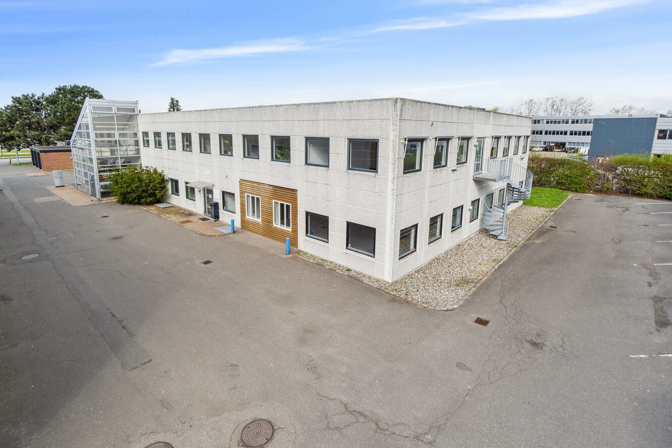 En rektangulær kontorbygning i to etager med mange vinduer, en udvendig trappe og et asfalteret område omkring under en delvist overskyet himmel.