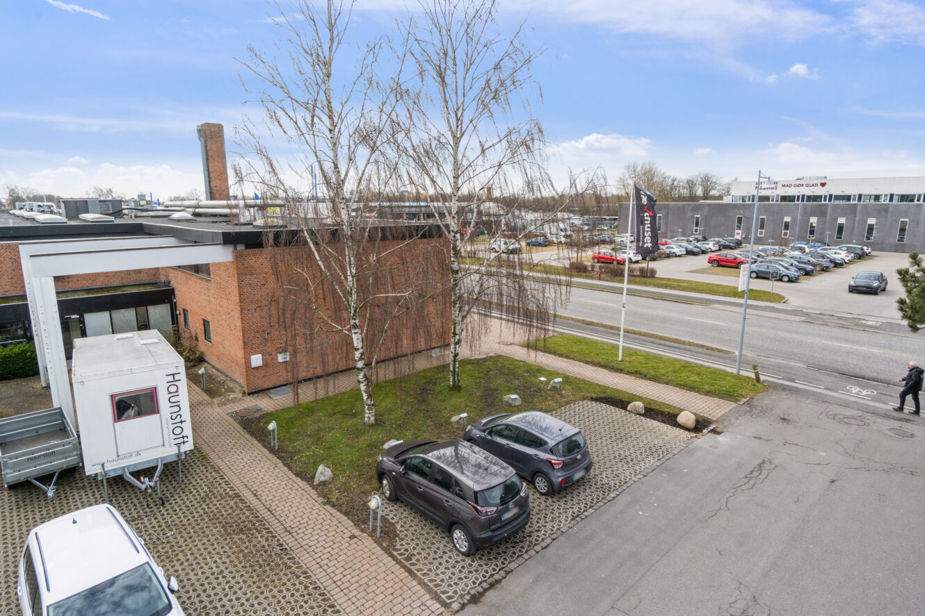 En asfalteret parkeringsplads med flere biler, en murstensbygning, nøgne træer og en vej med flere parkerede biler foran et industriområde.