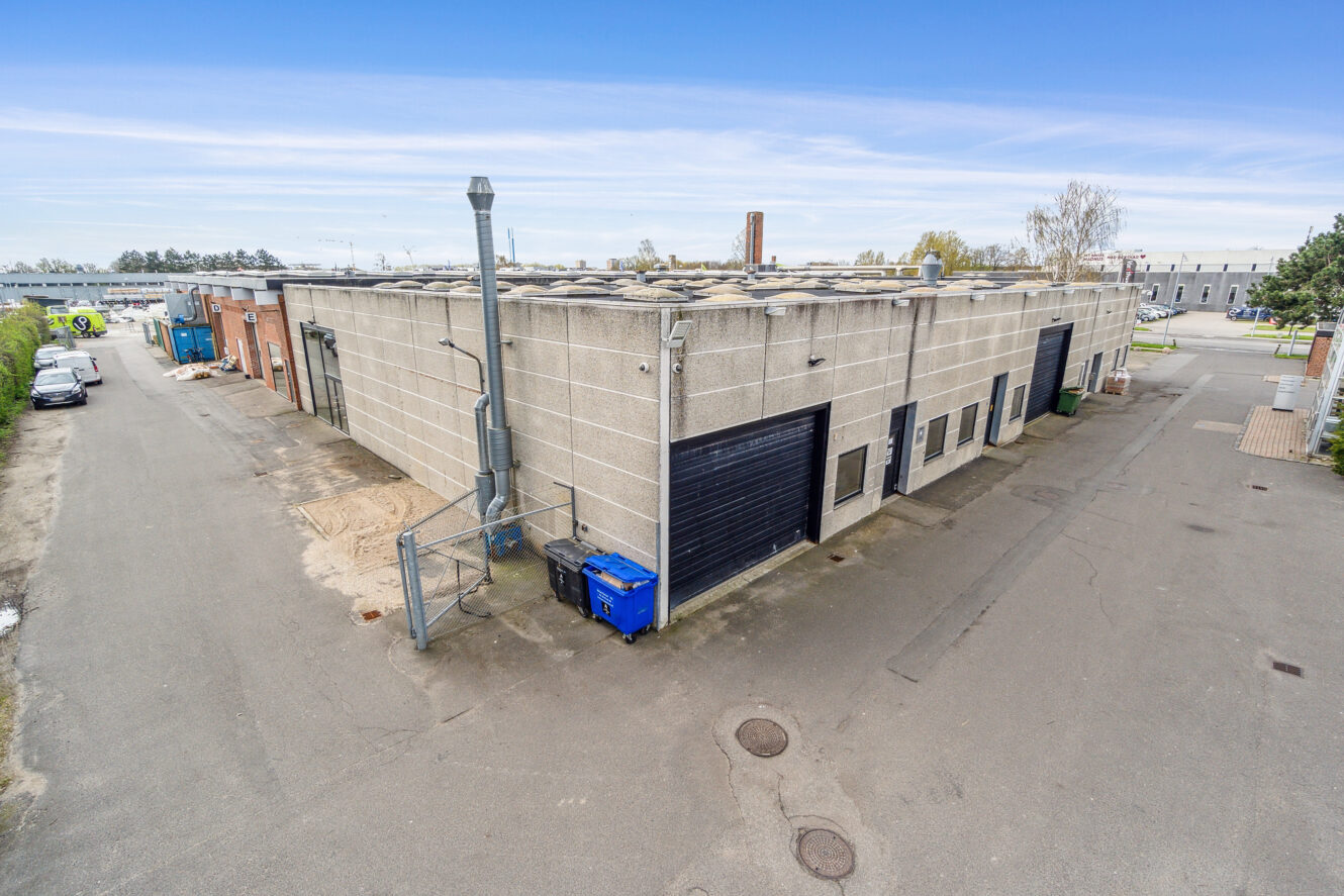 En stor, rektangulær industribygning med grå betonvægge, sorte garageporte og en blå affaldsspand står på en asfalteret grund under en klar himmel.