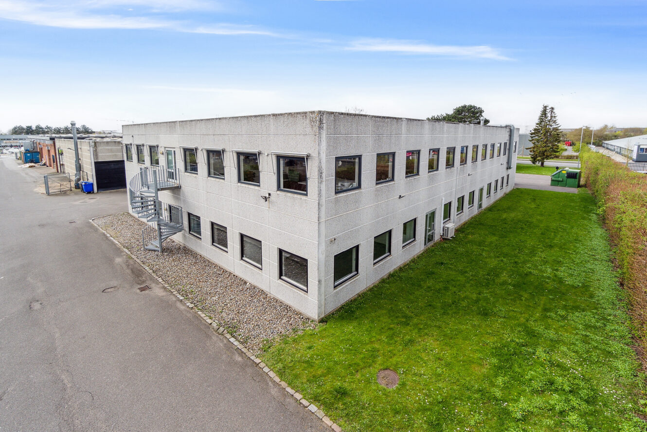 En rektangulær kontorbygning i to etager med grå beton udvendigt, mange vinduer og en vindeltrappe i metal. Bygningen er omgivet af græs og et asfalteret område.