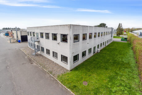 En rektangulær kontorbygning i to etager med grå beton udvendigt, mange vinduer og en vindeltrappe i metal. Bygningen er omgivet af græs og et asfalteret område.