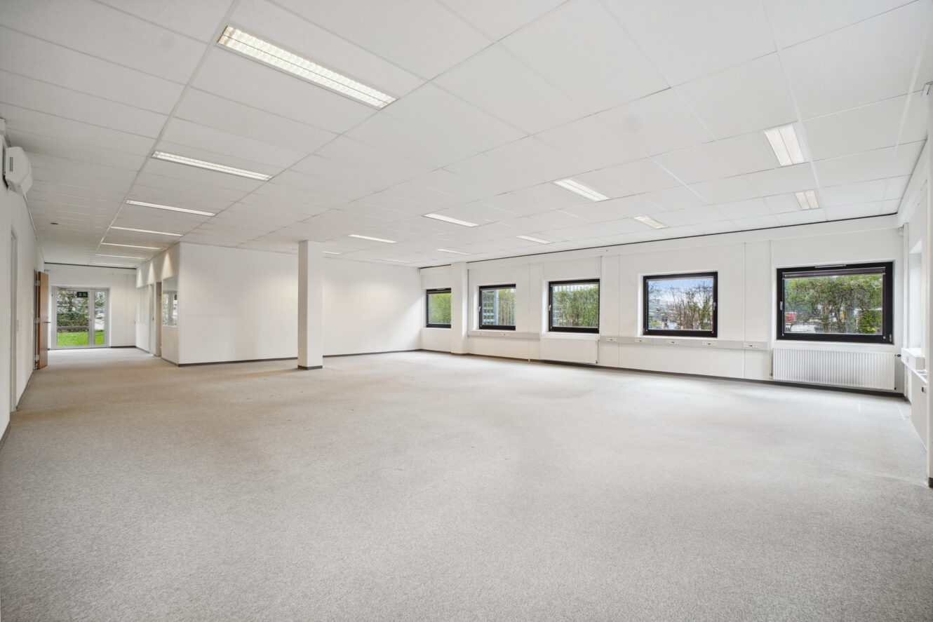 Rummeligt, tomt kontorlokale med hvide vægge, loftslamper, store vinduer og lysegråt gulvtæppe. Der kommer naturligt lys ind fra vinduer på to sider.