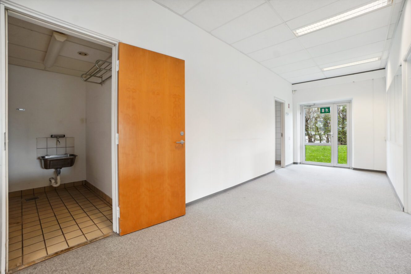 Et lille tomt rum med en brun dør, der åbner til et flisebelagt vaskeområde med en vask, hvide vægge, gulvtæppe og en glasdør, der fører ud i det fri.