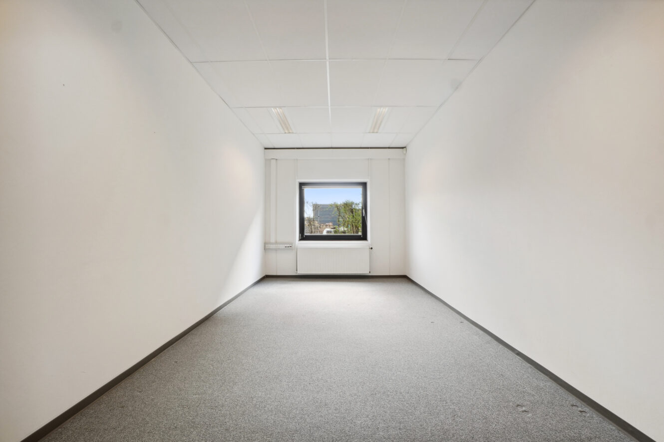 Et lille, tomt kontorlokale med hvide vægge, et gråt gulvtæppe og et enkelt vindue i den fjerneste ende, der lukker dagslys ind.