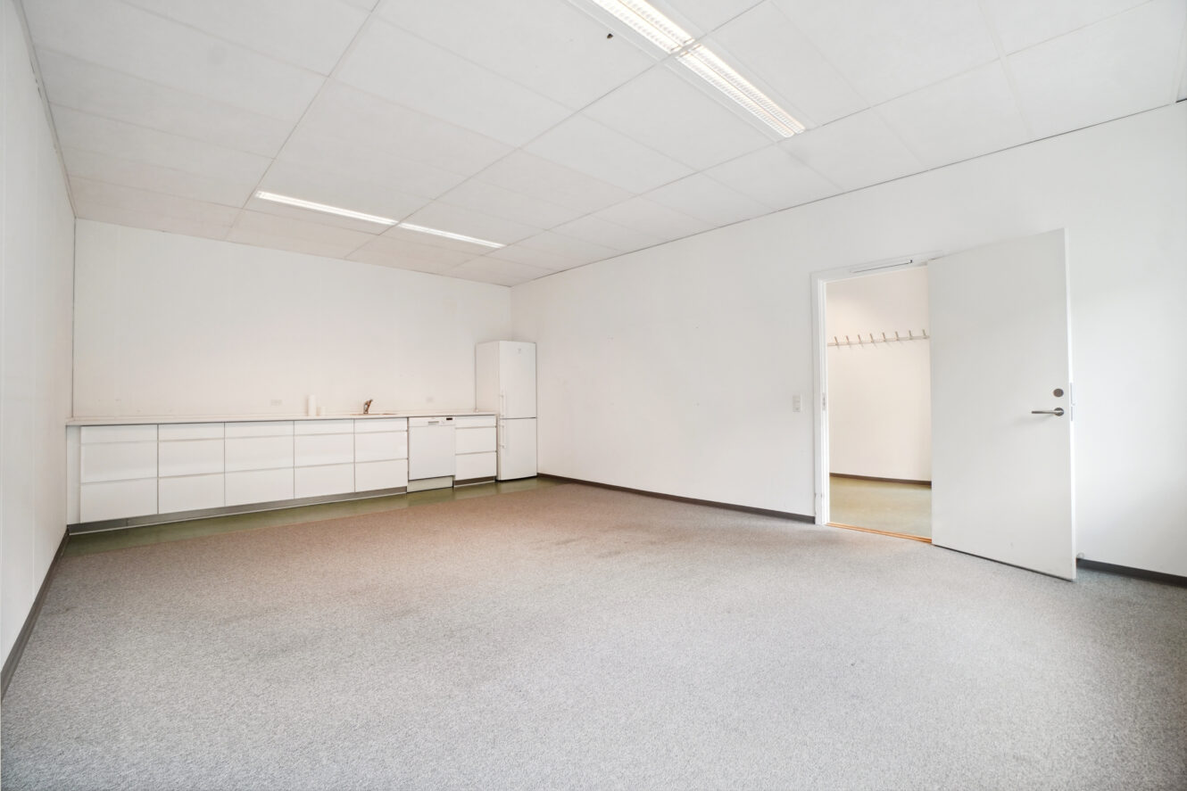 Et tomt rum med hvide vægge, gråt gulvtæppe, en række hvide skabe, et lille køleskab, en vask og en åben dør, der fører ud til en gang med knager.