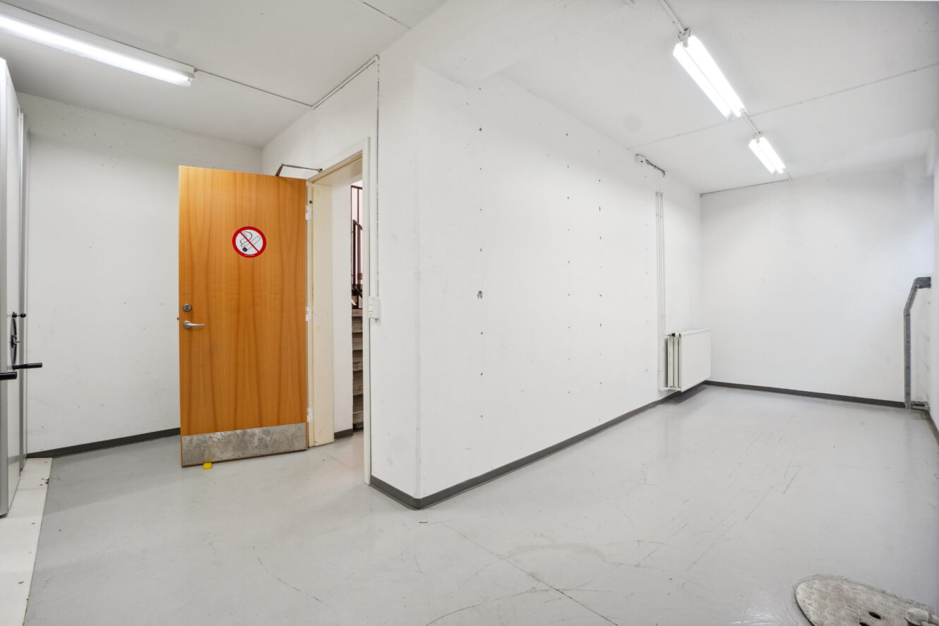 Tomt hvidt rum med lysstofrør, en åben trædør med et rygning forbudt-skilt og en radiator under et vindue på højre væg.