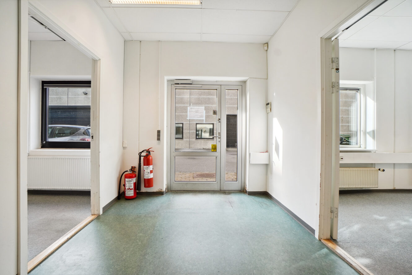 En gang med en glasdør, der fører ud, to åbne døråbninger på hver side, brandslukkere ved venstre væg og vinduer, der vender ud mod en stenbygning.