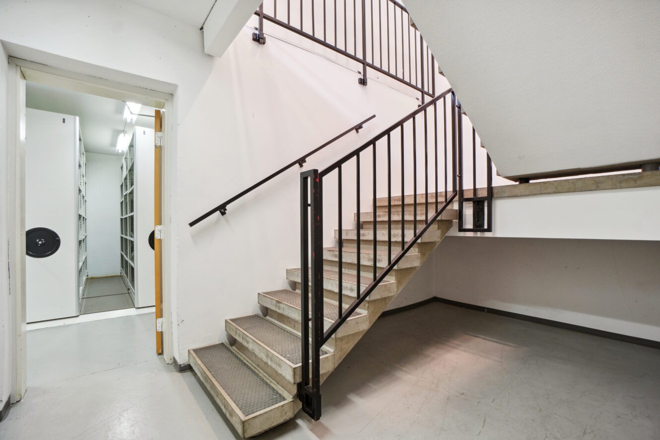 Betontrappe med metalgelænder i et rent indendørs område med hvide vægge; en døråbning til venstre fører til et rum med hylder og opbevaringsenheder.
