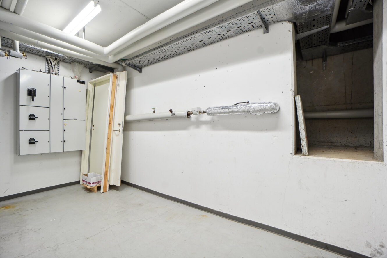 Et bryggers med elektriske paneler på venstre væg, synlige rør, en hvid dør og en åben rektangulær ventilations- eller adgangsskakt på højre side.