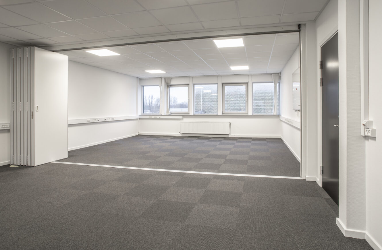 Tomt kontor eller konferencelokale med grå tæppefliser, hvide vægge, loftslamper, foldevæg og store vinduer, der lukker naturligt lys ind.