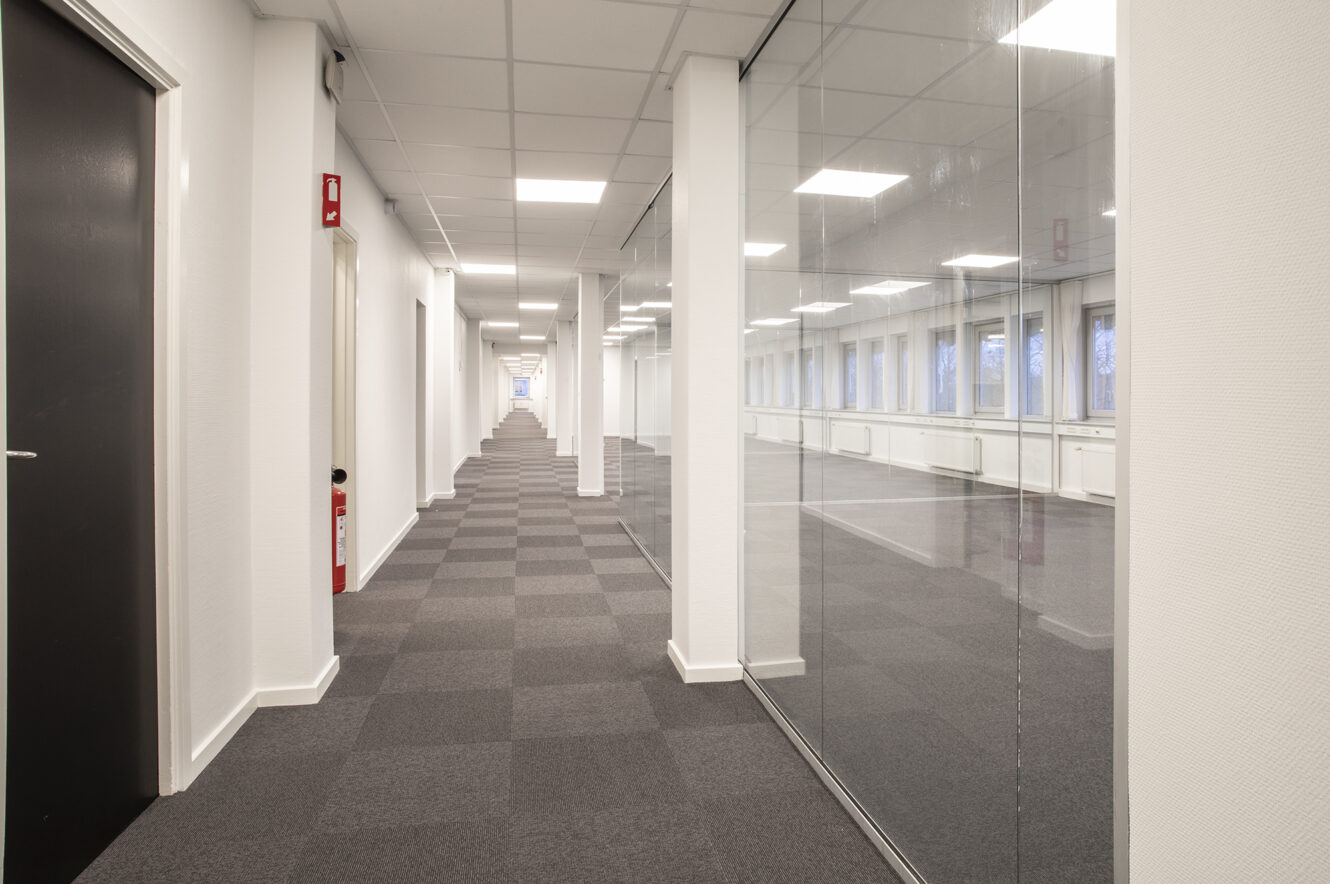 Lang kontorgang med gråternet tæppe, hvide vægge, glasskillevægge til højre og lukkede døre til venstre. Rummet er stærkt oplyst med loftslamper.