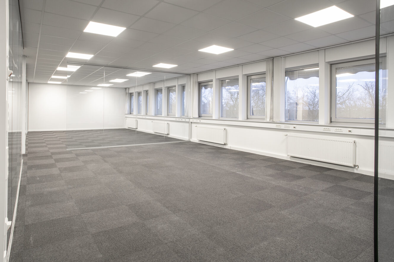 Tomt kontorlokale med gråt gulvtæppe, hvide vægge, store vinduer langs den ene side og lysstofrør i loftet.