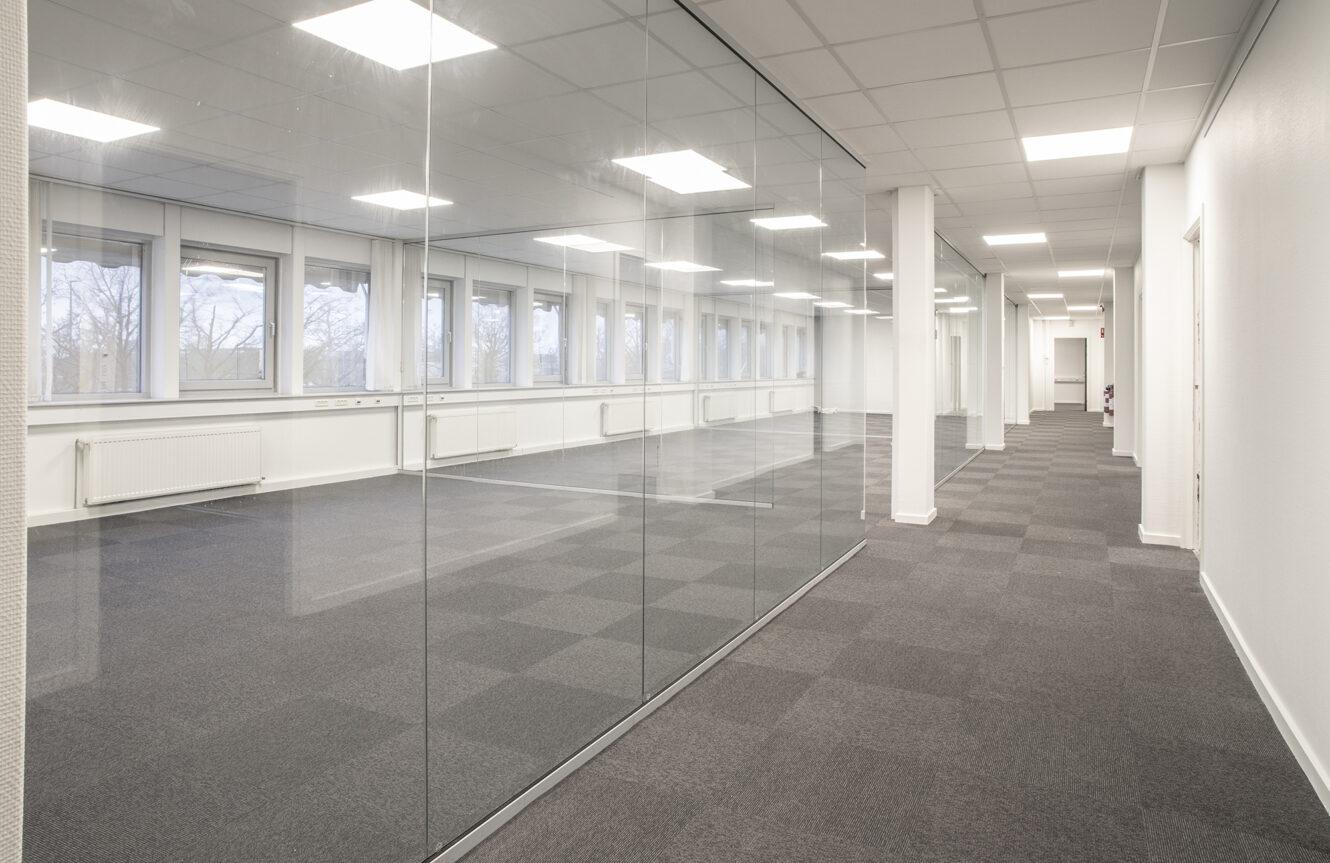 Tomme, moderne kontorlokaler med glasvægge, grå tæppefliser, hvide vægge og store vinduer, der lukker naturligt lys ind.