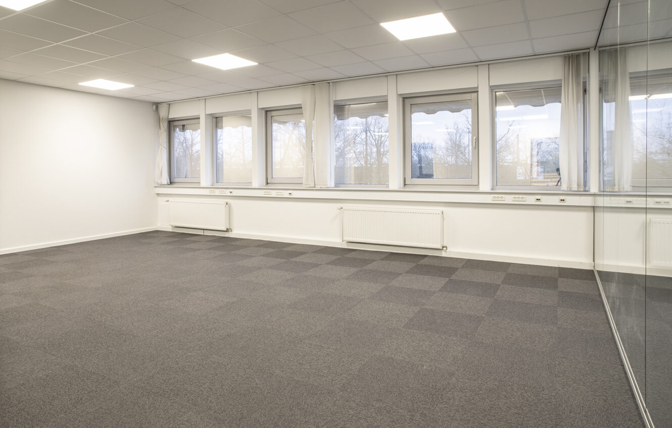 Tomt kontorlokale med gråt gulvtæppe, hvide vægge, en række vinduer, radiatorer og en glasskillevæg i højre side.