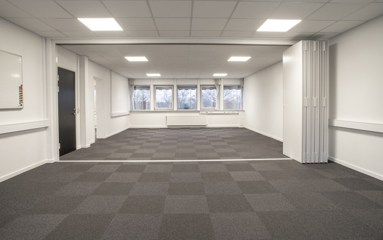 Et stort, tomt kontorlokale med gråternet gulvtæppe, hvide vægge, loftslamper, en sammenklappelig skillevæg, vinduer og en lukket sort dør.