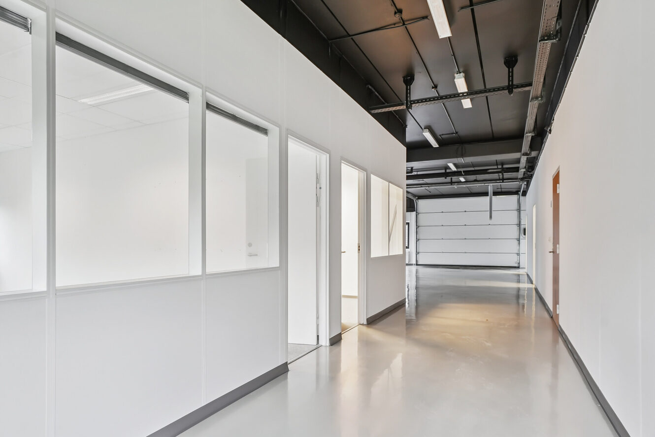 En moderne, tom gang med hvide vægge, kontorvinduer af glas, flere døre og et poleret gråt gulv under sorte loftsarmaturer.
