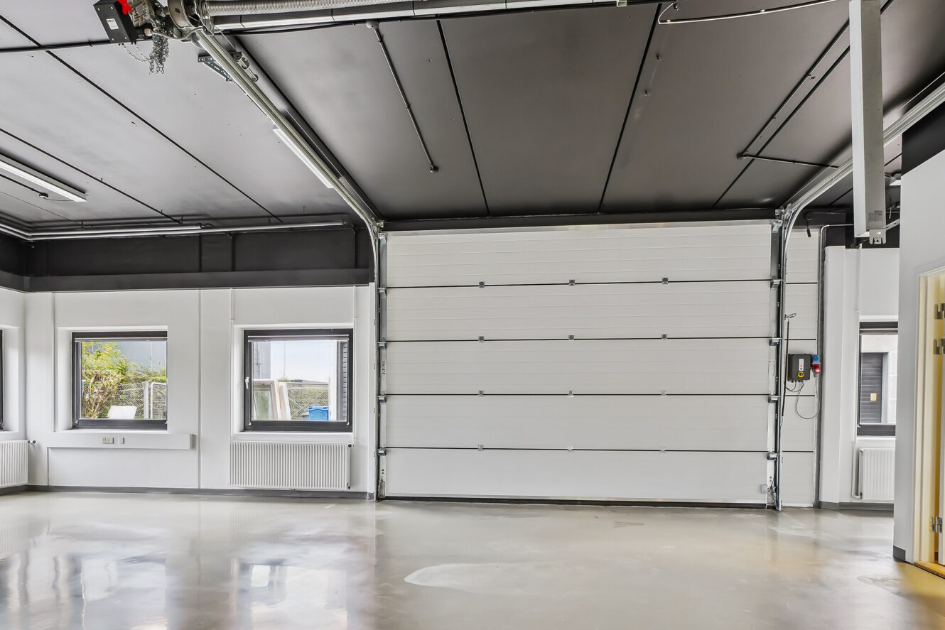 Tomt industrielt garageinteriør med en lukket hvid rulleport, poleret betongulv, store vinduer og synlige rør i loftet.