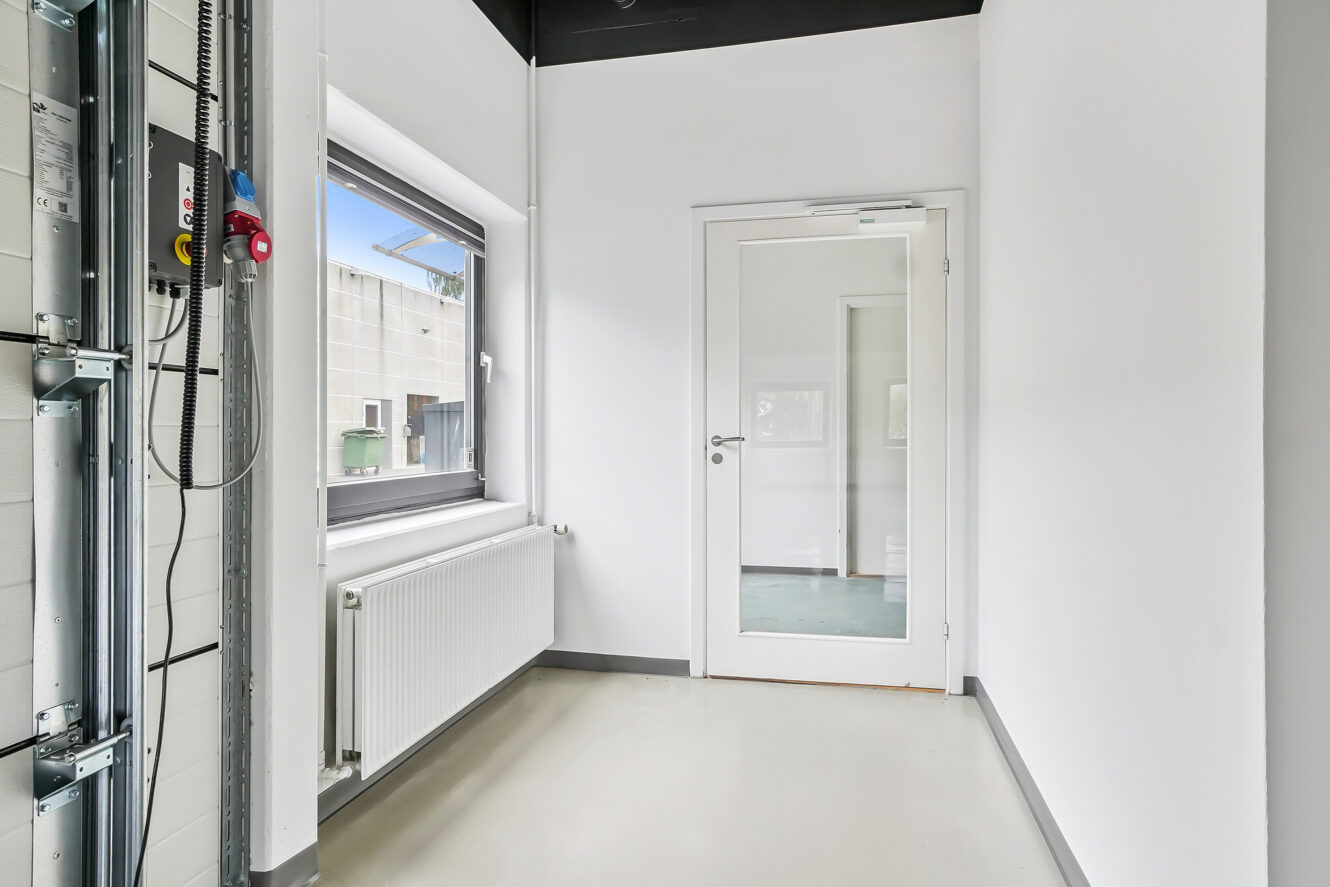 En lille, ren gang med hvide vægge, et vindue til venstre, en radiator nedenunder og en glasdør, der fører ind til et andet rum for enden.
