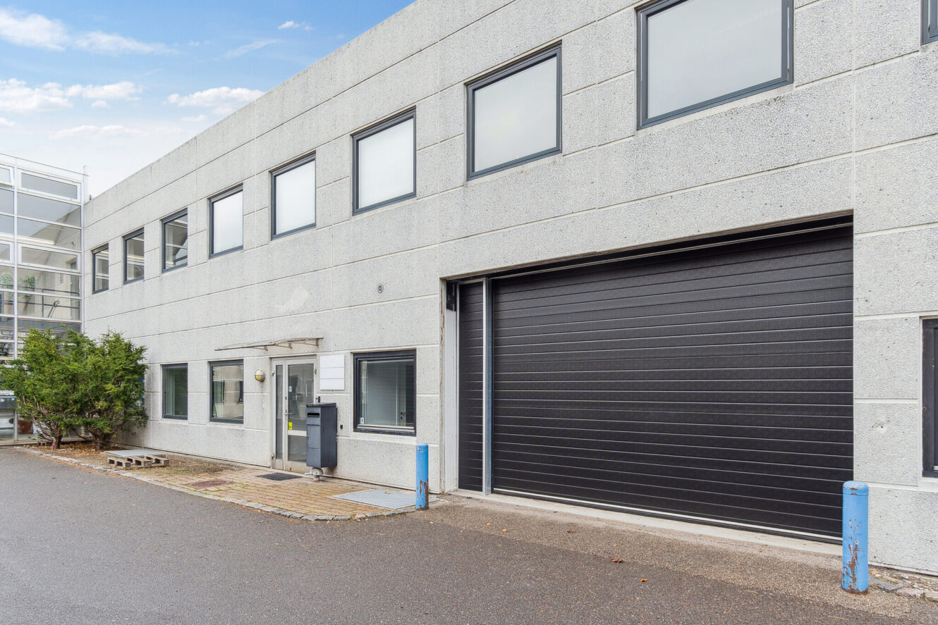 En moderne industribygning i to etager med betonvægge, sorte vinduer, en stor sort garageport og en indgangsdør af glas.