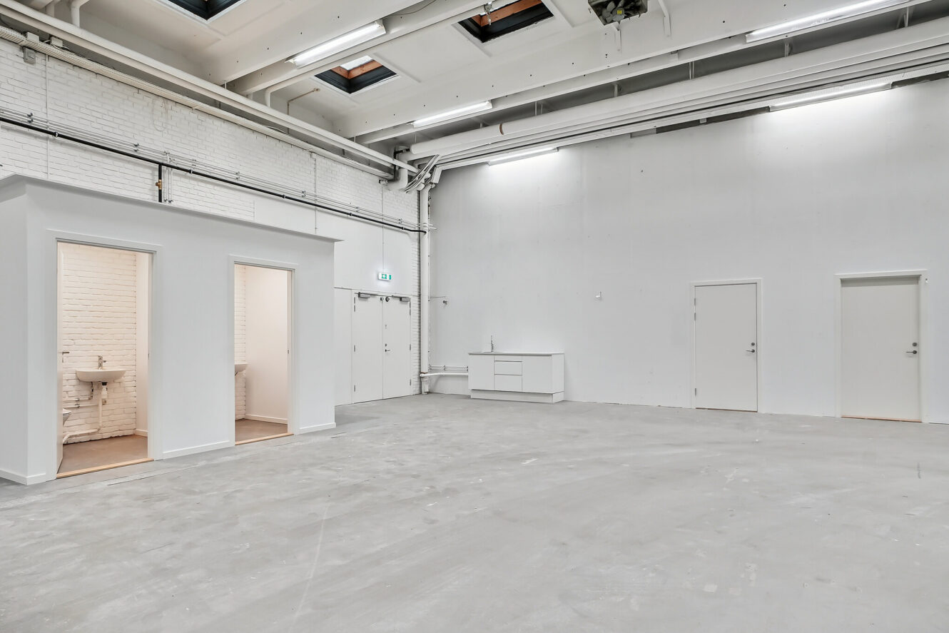 Stort, tomt industrilokale med hvide vægge, højt til loftet, ovenlys og tre døre; to rum med håndvaske er synlige på venstre side.