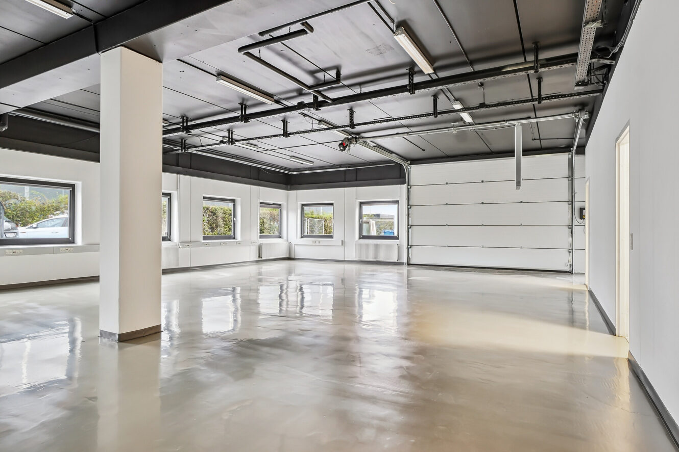 Rent, tomt industrilokale med poleret betongulv, hvide vægge, flere vinduer, synlige rør i loftet og en stor garageport.