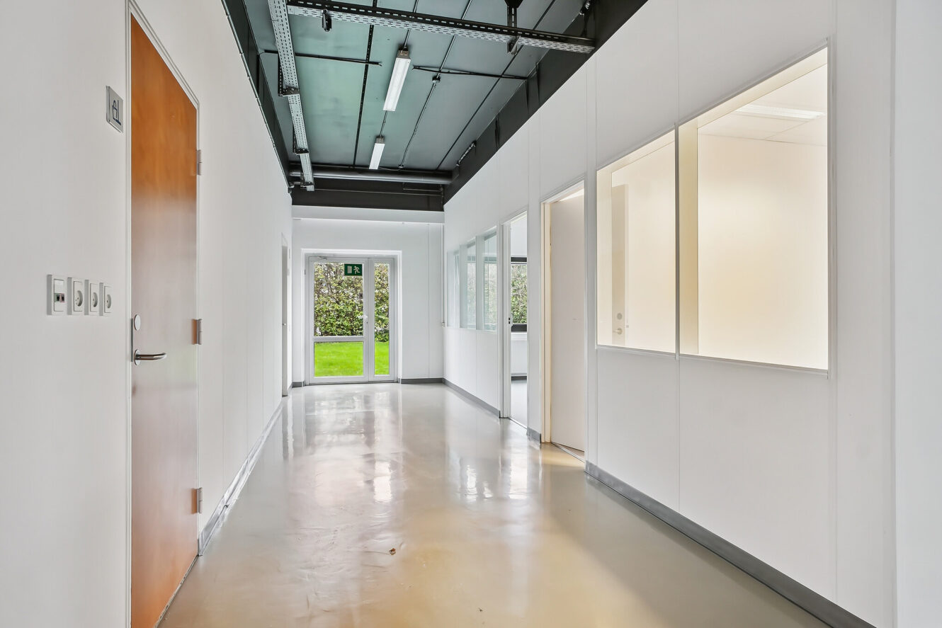Moderne gang med hvide vægge, kontorvinduer i glas til højre, en trædør til venstre og en dør for enden, der fører ud til et græsklædt udendørsareal.