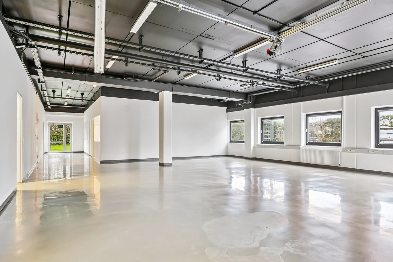 Rummeligt, tomt erhvervslokale med poleret betongulv, hvide vægge, store vinduer og synlige sorte loftsrør og armaturer. Naturligt lys kommer ind gennem flere vinduer.
