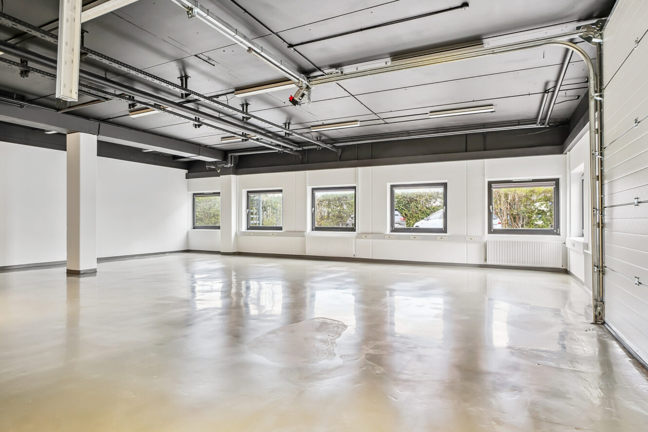 Rummeligt, tomt industrilokale med polerede betongulve, hvide vægge, store vinduer og synlige rør i loftet. Naturligt lys kommer ind gennem vinduerne.