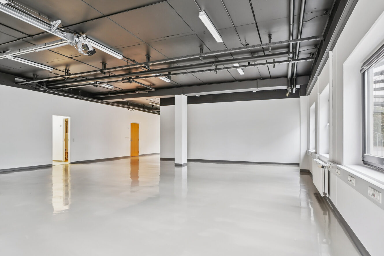 Et stort, tomt rum med polerede gulve, hvide vægge, store vinduer, synlige rør i loftet og fluorescerende belysning. En åben døråbning og en lukket trædør er synlige.