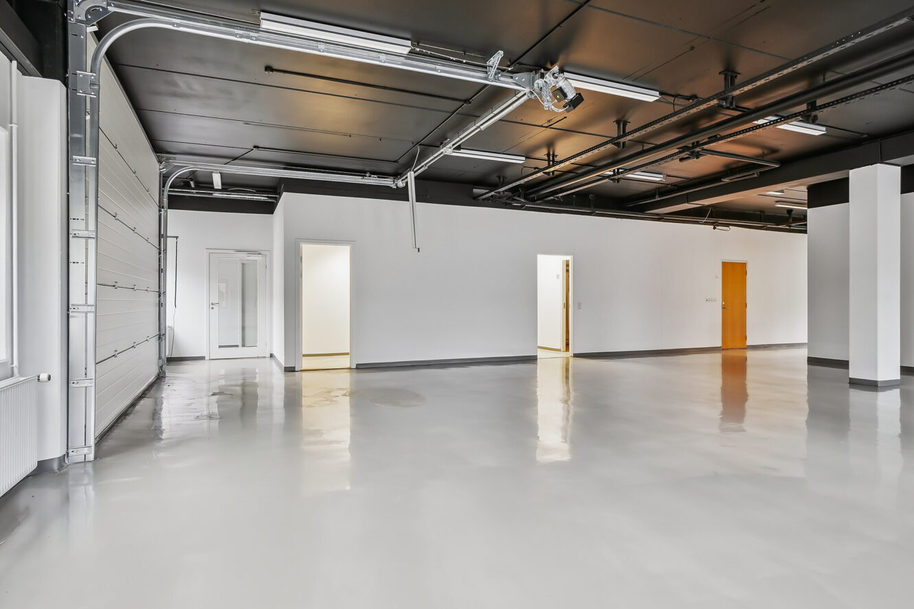 Stort, tomt industri- eller forretningslokale med poleret betongulv, hvide vægge, synlige rør i loftet og flere døråbninger og en garageport.