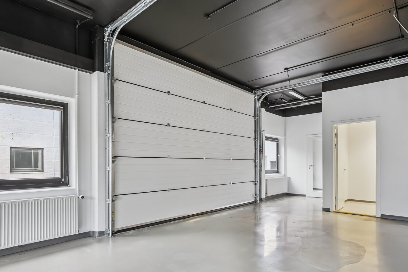 Interiørbillede af et tomt industriområde med en stor garageport, to vinduer, hvide vægge, poleret gulv og en åben døråbning, der fører til et andet rum.