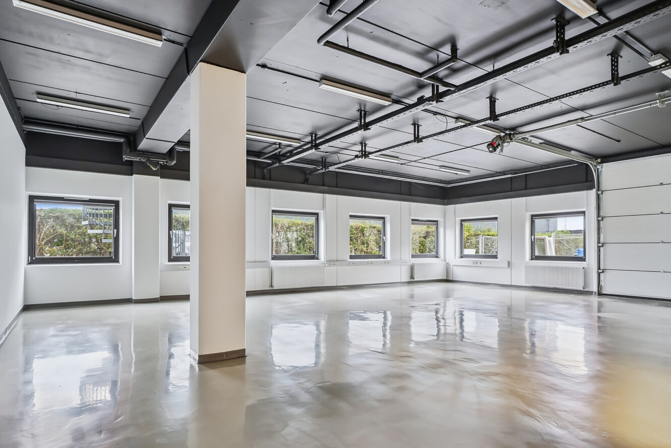 Rummeligt, tomt forretningslokale med poleret betongulv, hvide vægge, flere vinduer og et loft med synlige armaturer og belysning.