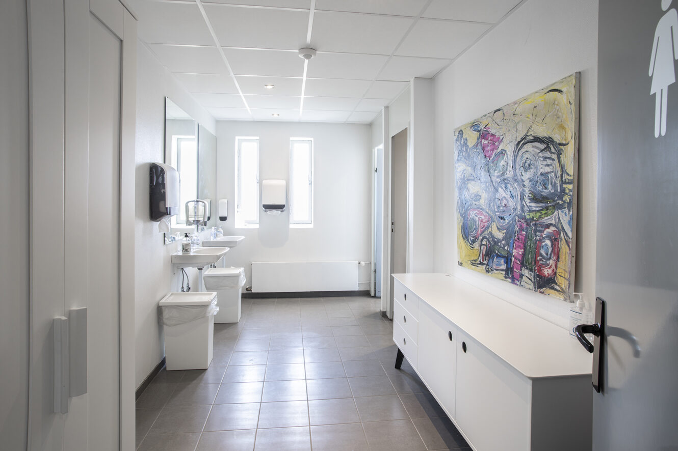 Et moderne dametoilet med hvide vægge, håndvaske, spejle, en skraldespand, abstrakt vægkunst og naturligt lys fra to smalle vinduer.